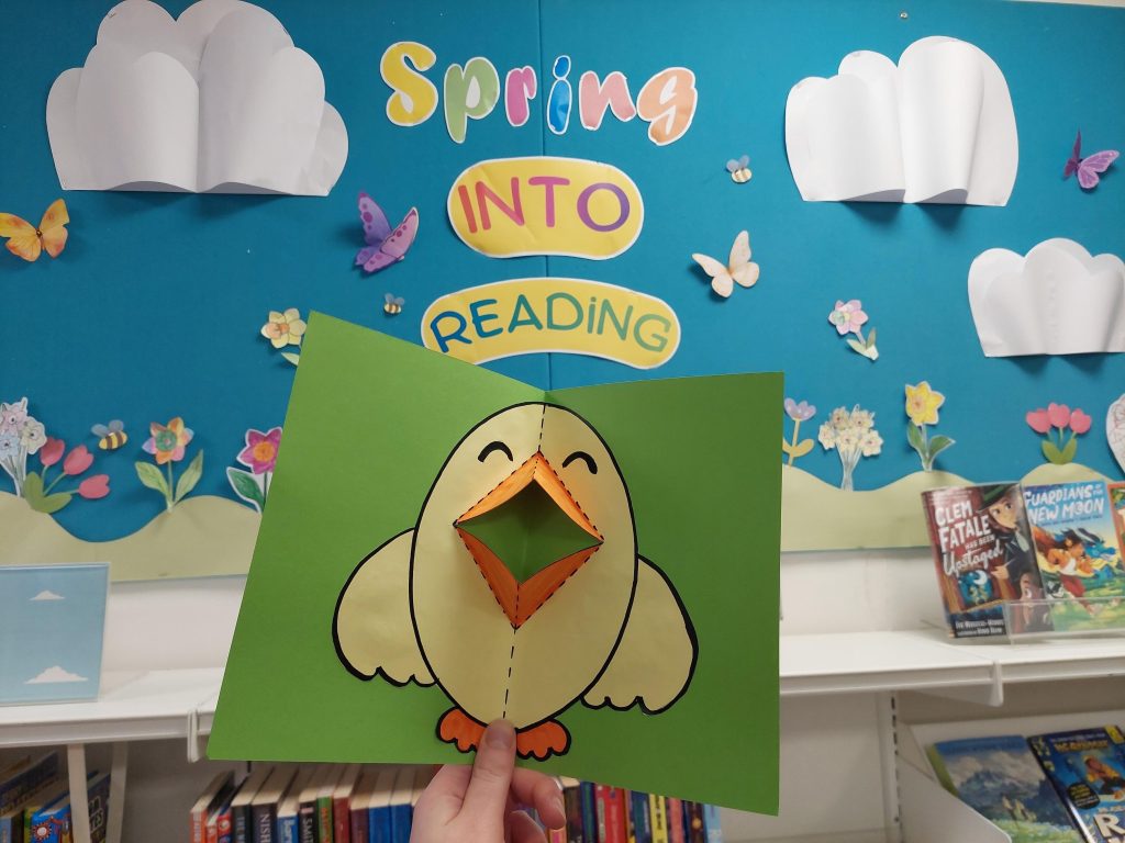 A colourful library display themed “Spring into Reading.” The backdrop is a bright blue board decorated with paper clouds, butterflies, bees, and spring flowers. At the top, large playful cut-out letters read “Spring INTO READING,” with “Spring” in multicoloured letters and “INTO READING” on yellow shapes beneath. In the foreground, a hand holds up a green piece of card featuring a simple, cheerful chick illustration. The chick has a folded, 3D orange paper beak that pops out from the page, giving it a playful, interactive look. Behind the display, bookshelves are visible with children’s books, including titles such as “Clem Fatale Has Been Betrayed” and “Guardians of the New Moon,” reinforcing the reading theme.