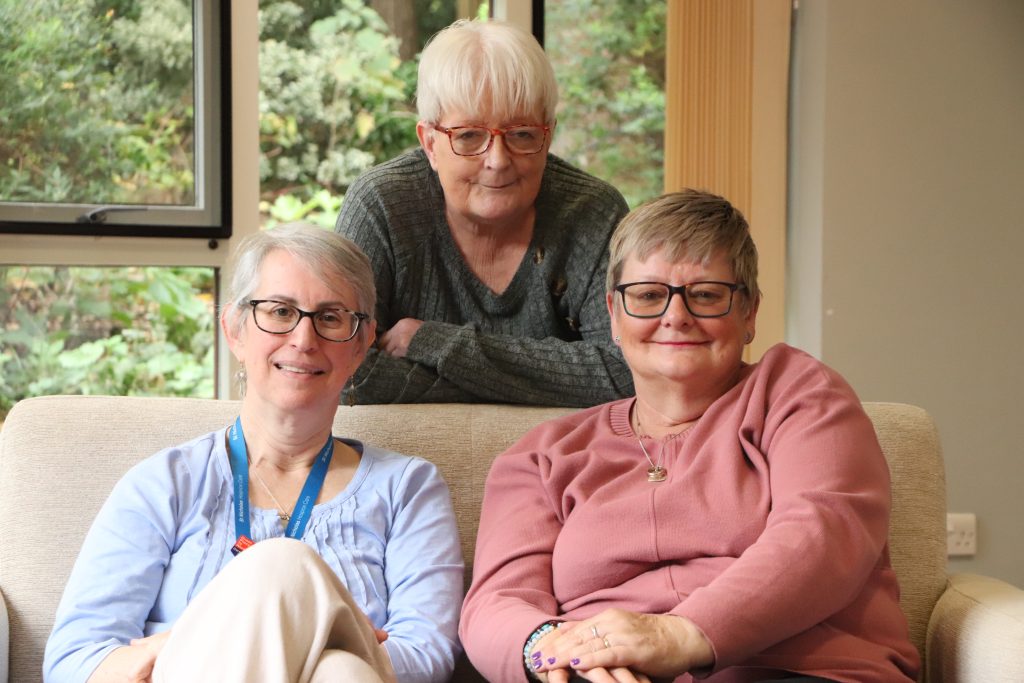 A group of three women sit together indoors in a lounge area, smiling towards the camera. Two women sit on a cream sofa while a third leans over the back of it. Large windows behind them look out onto greenery and trees outside.