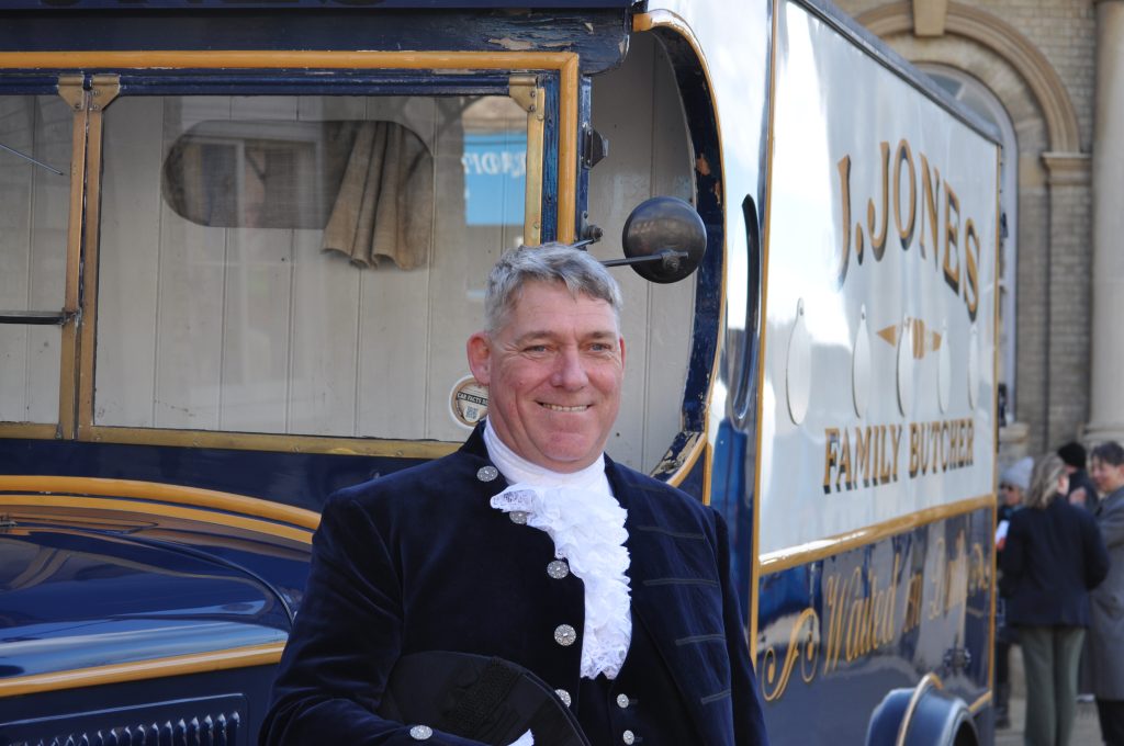 A man in traditional formal attire, including a dark coat with decorative buttons and a white cravat, stands smiling beside a vintage delivery van. The van is painted in cream and blue and reads “J. Jones Family Butcher” on the side, with people and a building visible in the background.