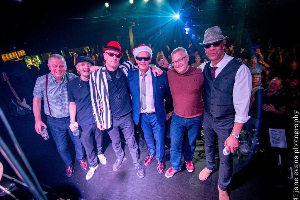 A group of six men stand together on a stage, posing arm-in-arm in front of a lively audience. They are dressed in a mix of casual and retro-inspired outfits, including hats, sunglasses, a striped blazer, and a Santa hat on one member, suggesting a themed or festive performance.The stage lighting casts a blue glow, and the audience behind them has their hands raised, indicating an energetic live music atmosphere. Musical equipment, including a guitar and stage monitors, is visible around them.Along the right edge of the image is a vertical watermark reading “© jane evans photography”.