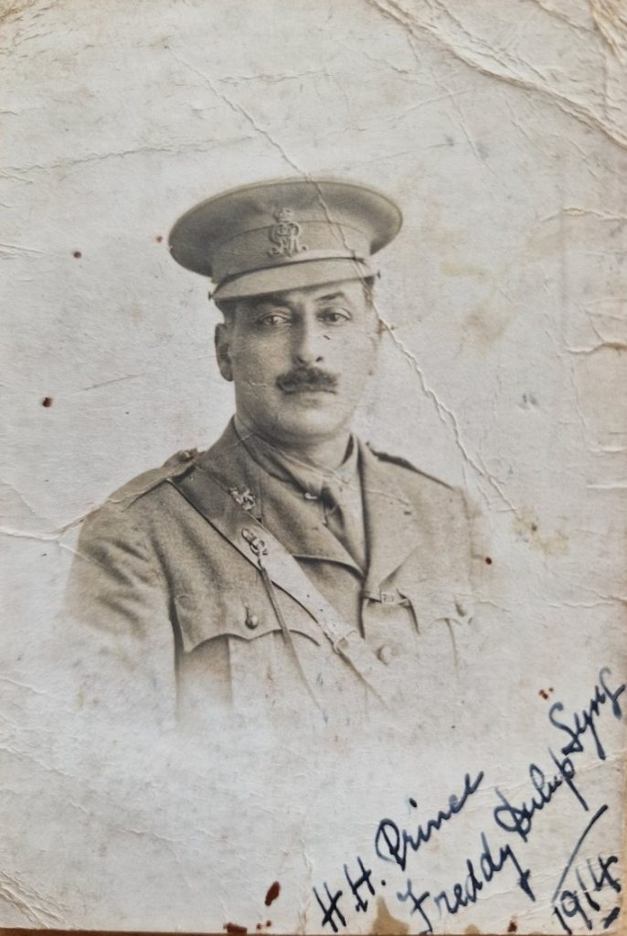 An aged black-and-white portrait photograph of a man in early 20th-century military uniform. He is shown from the chest up, facing forward with a neutral expression and a neatly groomed moustache. He wears a peaked military cap with an insignia on the front, along with a buttoned tunic and a strap crossing diagonally over his chest.The photograph shows visible signs of age, including creases, stains, and slight discoloration across the surface. In the lower right corner, there is a handwritten inscription in ink reading: “H.H. Prince Frederick Duleep Singh 1914,” written at an angle.The background is plain and faded, placing full focus on the subject and the historic nature of the image.