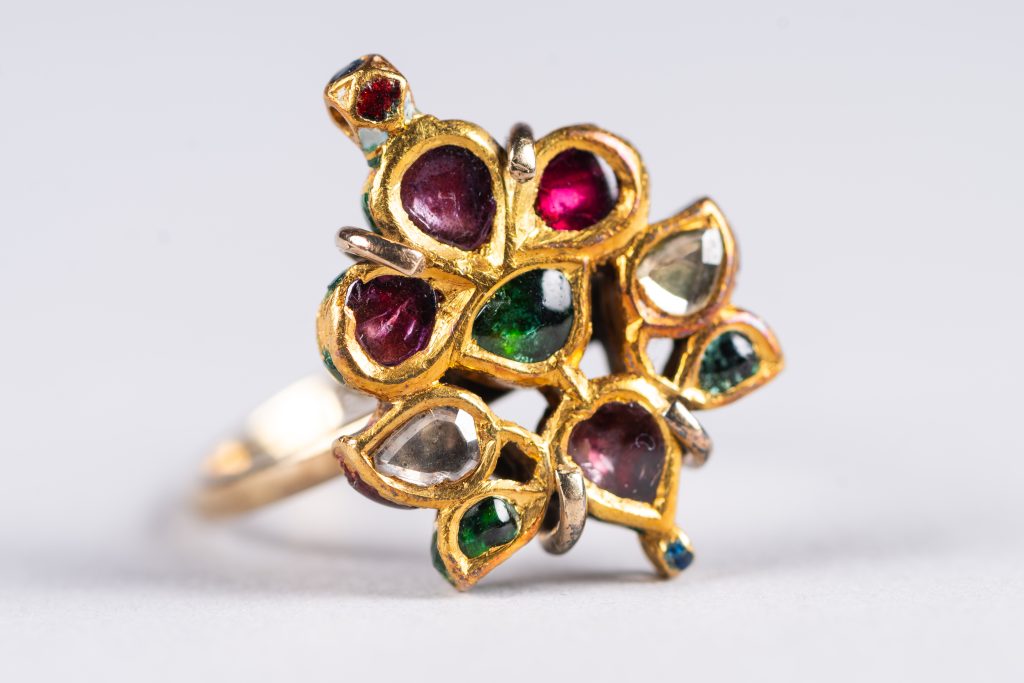 A close-up photograph of an ornate gold ring set with multiple colourful gemstones. The ring features an intricate cluster design, with teardrop-shaped settings arranged like petals around a central point.The gemstones include deep red, green, and clear stones, each set in raised gold bezels. The gold has a slightly textured, handcrafted appearance, and the stones vary slightly in size and shape, giving the piece an organic, decorative look.The ring is photographed against a plain light grey background, with soft lighting highlighting the reflective surfaces of the gold and the rich colours of the gemstones.