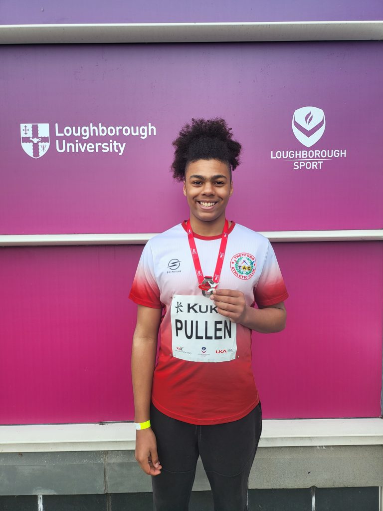 A young athlete stands smiling in front of a pink backdrop displaying “Loughborough University” and “Loughborough Sport” logos. They are wearing a red and grey Thetford Athletic Club shirt and holding up a silver medal on a red ribbon. A competition bib on their chest reads “PULLEN,” and a green wristband is visible on their wrist.