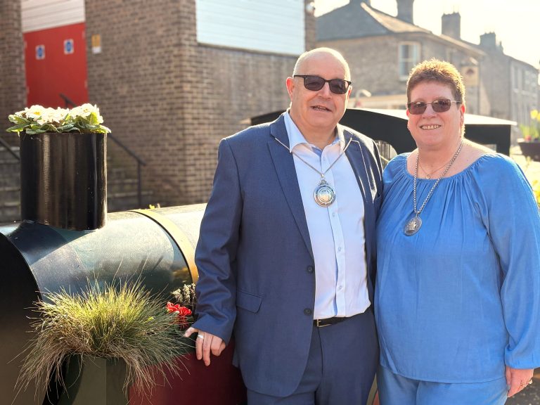 A man and woman stand together outdoors in bright sunlight, posing beside a decorative black metal structure with planted flowers. The man, wearing a blue suit jacket, light shirt, and dark sunglasses, has a ceremonial chain of office around his neck. The woman, standing beside him, wears a loose blue top, glasses, and a similar ceremonial medallion. Both are smiling and facing the camera. Behind them are brick buildings, including one with a red door, suggesting a town centre or civic setting. The scene has a warm, formal yet friendly feel, indicating a civic role or official appointment.