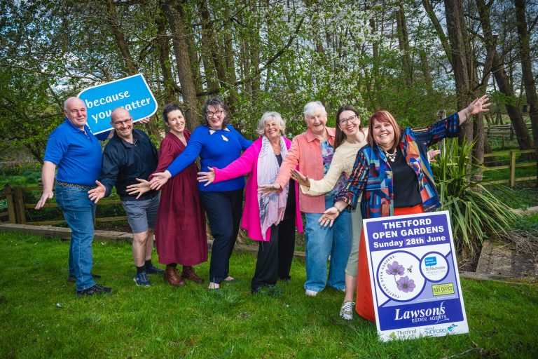A group of volunteers and organisers pose outdoors beside a sign promoting Thetford Open Gardens on Sunday 28th June in aid of St Nicholas Hospice Care. The group stands on grass with trees and woodland in the background, many holding their arms out and smiling towards the camera. Signs reading “because you matter” and the event board are visible.