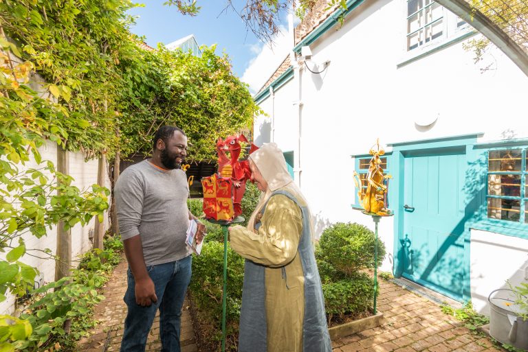 Man speaking with a costumed guide in a sunlit courtyard garden outside a white building with turquoise doors and windows. Decorative dragon sculptures on poles stand between them, while leafy plants and brick pathways surround the scene.