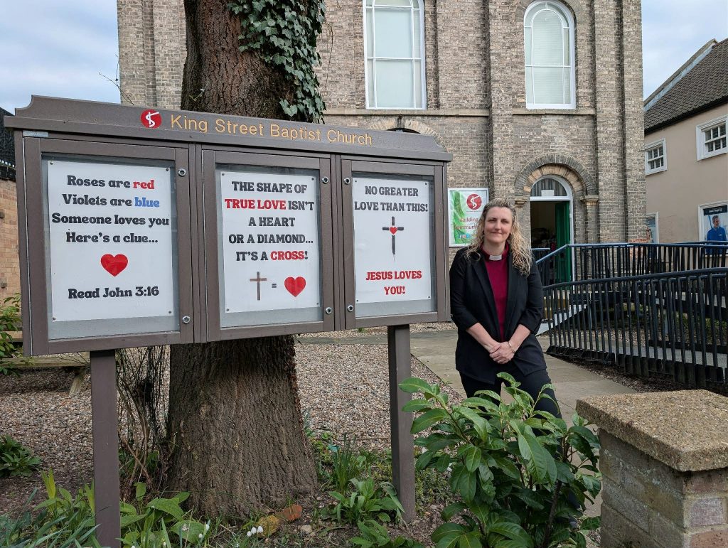 A woman stands outside King Street Baptist Church beside a three-panel noticeboard mounted in front of a tree. The church building, made of light-coloured brick with tall arched windows, is visible behind her, along with a ramp and railings leading to the entrance. The woman, wearing a black jacket over a red top, stands with her hands clasped and smiles towards the camera. The noticeboard header reads “King Street Baptist Church” and displays three posters with Christian messages. The left poster reads: “Roses are red Violets are blue Someone loves you Here’s a clue…” with a red heart icon, followed by: “Read John 3:16” The centre poster reads: “THE SHAPE OF TRUE LOVE ISN’T A HEART OR A DIAMOND… IT’S A CROSS!” with a cross symbol and a heart icon. The right poster reads: “NO GREATER LOVE THAN THIS!” with a cross and heart symbol, followed by: “JESUS LOVES YOU!” Plants and greenery surround the base of the sign, adding to the outdoor setting.