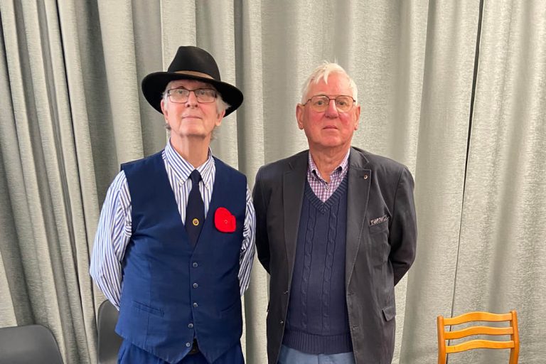 Photo of two older men standing side by side in front of grey curtains indoors. The man on the left is wearing a black hat, glasses, a striped shirt with tie, and a blue waistcoat with a red poppy pin. The man on the right is wearing glasses, a dark blazer over a navy jumper and checked shirt. An empty chair and part of a table are visible at the edges of the image.