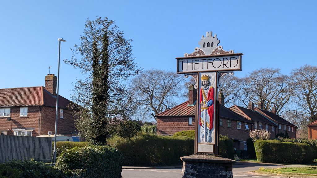 A roadside welcome sign for Thetford stands on a small stone base at a residential junction. The sign features a decorative painted panel of a crowned historical figure wearing a red cloak and holding a staff or sword, positioned beneath a white signboard with bold lettering reading “THETFORD”. Behind the sign are rows of red-brick houses with tiled roofs, neatly trimmed hedges, and a quiet street curving away to the right. A tall, leafless tree covered in ivy stands to the left of the sign, alongside a modern streetlamp. The scene is brightly lit by clear sunshine under a blue sky, giving a crisp, calm suburban feel to the entrance of the town.