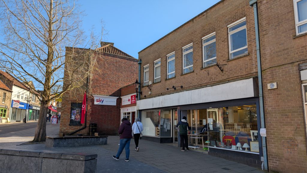 Street scene in Thetford town centre showing a row of brick buildings. A Sky and Vodafone shop sits on the corner, with a large empty retail unit next door displaying items in the window. A few pedestrians walk past, and a leafless tree stands in a paved seating area in the foreground under a clear blue sky.