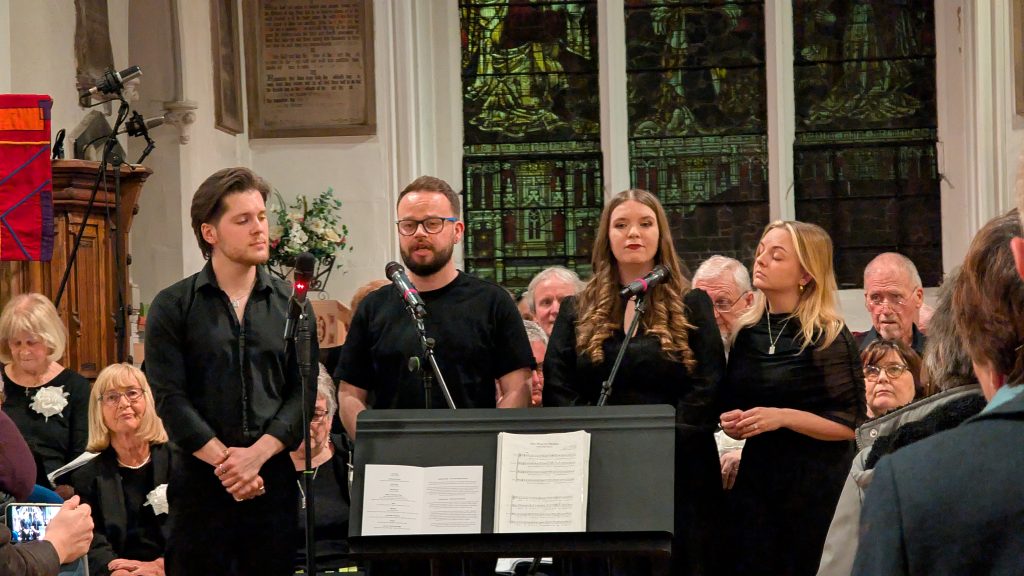 Four soloists from Thetford Singers perform at the front of St Cuthbert’s Church, standing behind microphones and a music stand, with members of the choir seated behind them. The singers are dressed in black, with a stained-glass window and church interior visible in the background, and audience members in the foreground watching the performance.