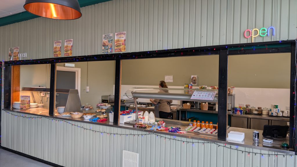 Interior of a café or food counter with a long serving hatch. A staff member works behind the counter, with hot food displays, cakes, snacks and drinks arranged along the front. Menu posters are mounted above, and an illuminated “open” sign and string lights decorate the serving area.