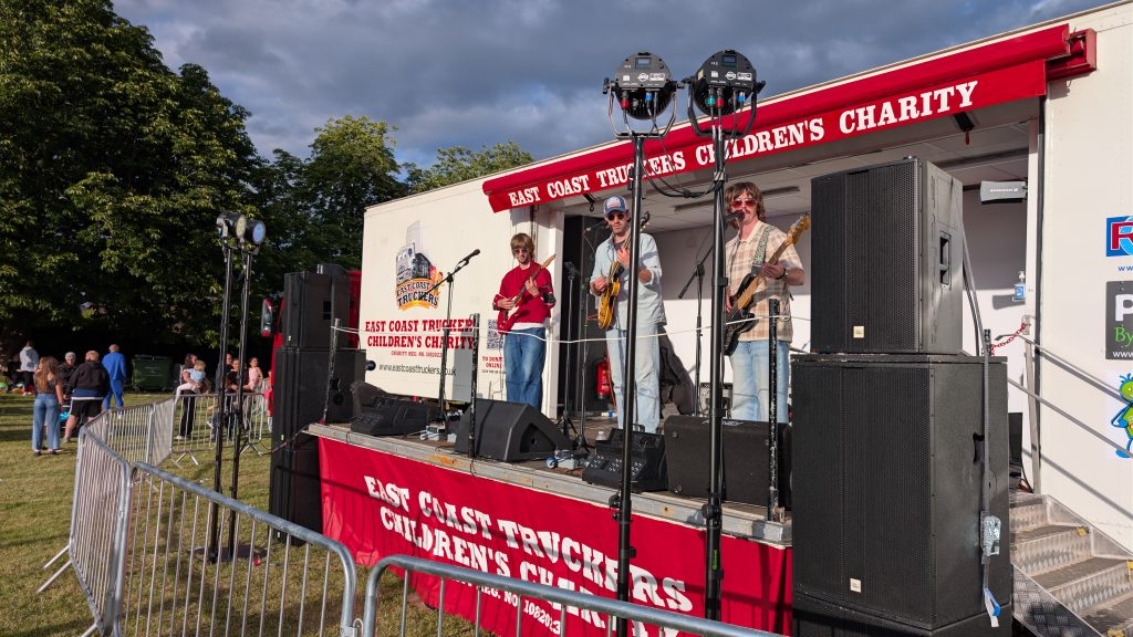 A live band performs on an outdoor stage mounted on a lorry operated by the East Coast Truckers Children's Charity. Three musicians play guitars and sing into microphones while large speakers and stage lighting frame the performance. People gather behind crowd barriers to watch beneath dramatic evening skies.