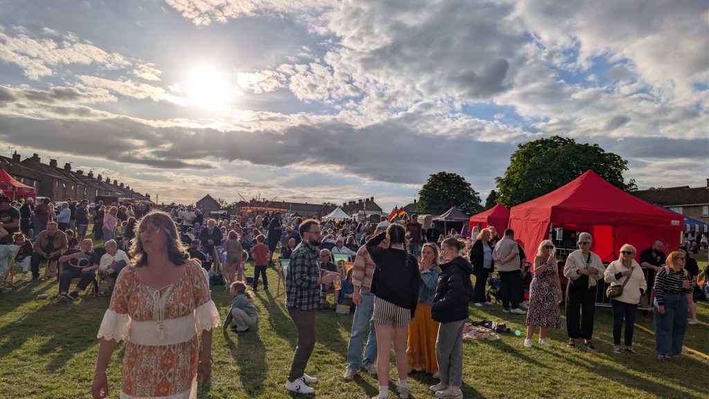 A large crowd enjoys an outdoor community festival on a grassy field under bright evening sunshine. Families and groups of friends relax on deckchairs, chat and queue at stalls beneath colourful gazebos, while entertainment continues in the background under a sky filled with dramatic clouds.