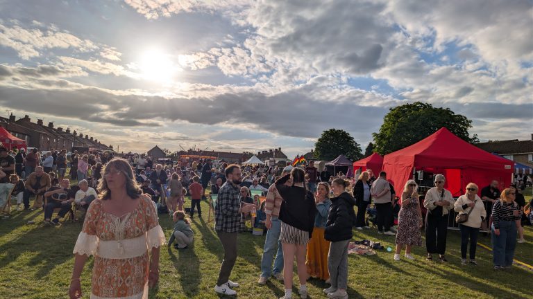 A large crowd enjoys an outdoor community festival on a grassy field under bright evening sunshine. Families and groups of friends relax on deckchairs, chat and queue at stalls beneath colourful gazebos, while entertainment continues in the background under a sky filled with dramatic clouds.