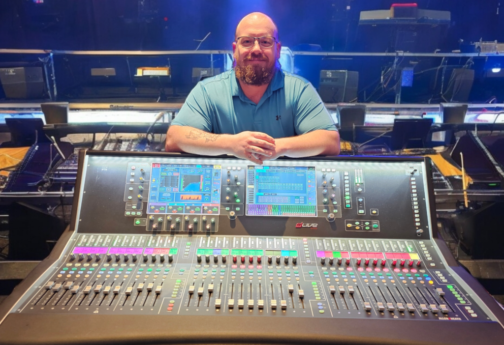 A man stands behind a large professional audio mixing desk in a dimly lit venue or theatre setting. He is smiling and leaning forward with his arms resting on the console. He has a shaved head, a full beard, and is wearing glasses and a blue polo shirt. The mixing desk in front of him is covered in rows of sliders, knobs, and brightly coloured buttons, with two illuminated digital screens displaying audio levels and controls. The brand name “dLive” is visible on the console. Behind him, the stage area is visible with speakers, cables, and lighting equipment under blue stage lighting, suggesting a live music or performance environment.