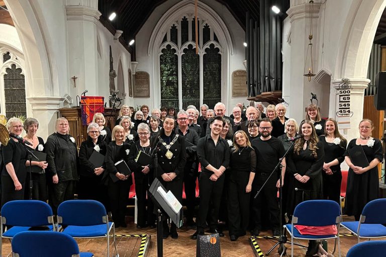 Thetford Singers choir members and soloists pose together inside St Cuthbert’s Church following their Spring concert. The group, dressed in black, stand in front of the altar beneath a large stained-glass window, with microphones and music stands visible in the foreground and rows of blue chairs set out for the audience.