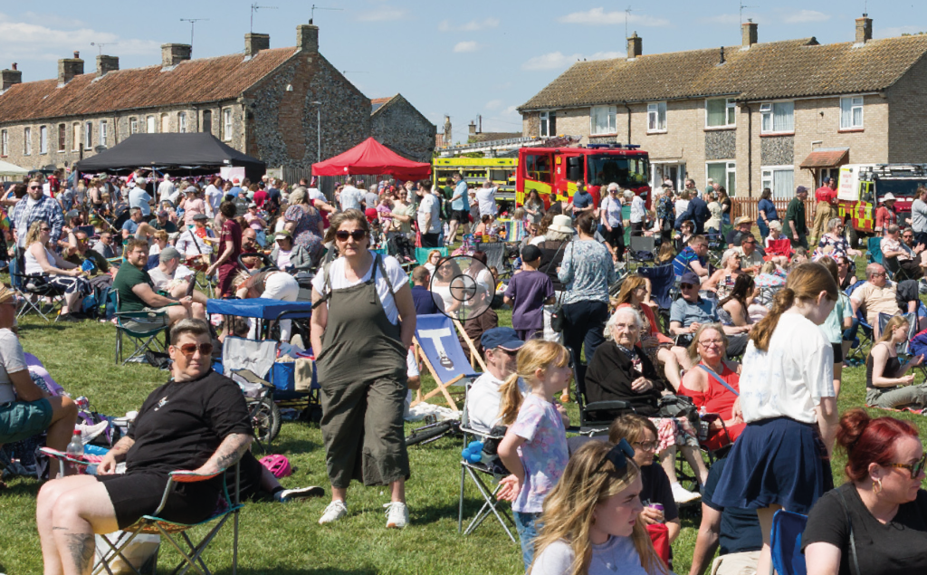 A large outdoor community event on a sunny day, with crowds of people sitting on camping chairs and blankets across a grassy field. Families and groups mingle, with children and adults moving through the scene. In the background are rows of houses, a red marquee tent, and a fire engine, suggesting a local fete or community gathering.