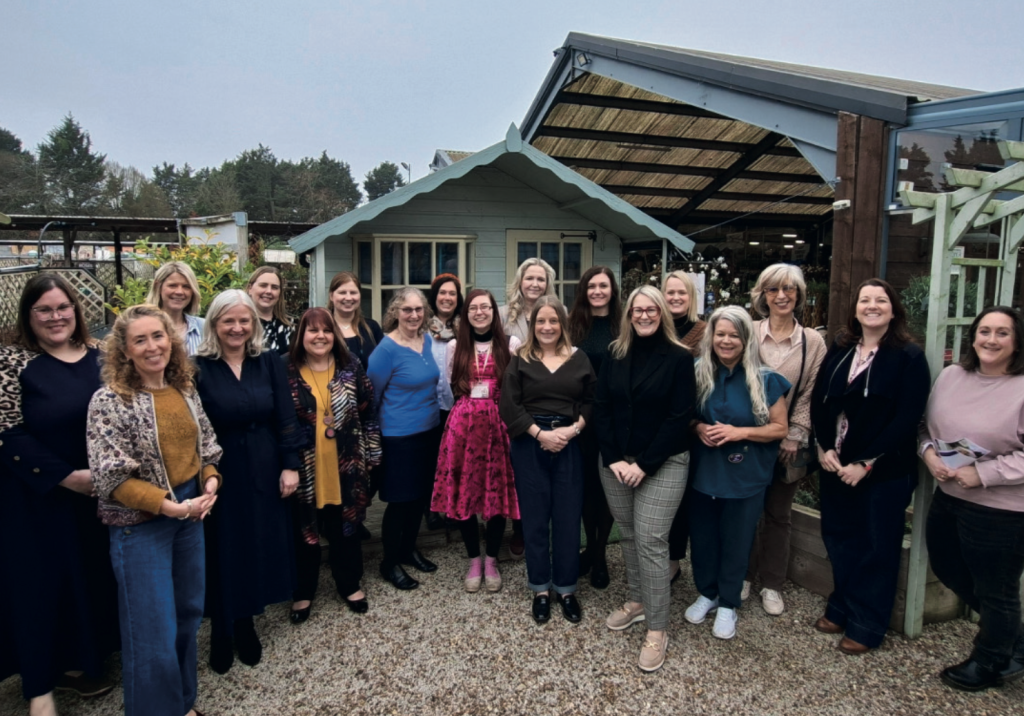 A large group of around twenty women stand together outdoors in front of a small garden-style building and plant nursery structures. They are arranged in a loose semi-circle, facing the camera and smiling. The women are dressed in a mix of casual and smart-casual clothing, including dresses, blouses, cardigans, and trousers. One woman near the centre stands out wearing a bright pink patterned dress with black tights and pink boots, while others wear more neutral tones such as black, navy, and earth colours. Behind them is a small painted wooden shed with a pitched roof and windows, flanked by larger open-sided garden structures with timber beams and roofing. Plants and garden displays are visible around the area, suggesting a garden centre or outdoor retail space. The ground is covered with light-coloured gravel, and the background includes trees and an overcast sky, giving a soft, muted lighting to the scene. The overall image suggests a group visit, team gathering, or community event in a garden or horticultural setting.
