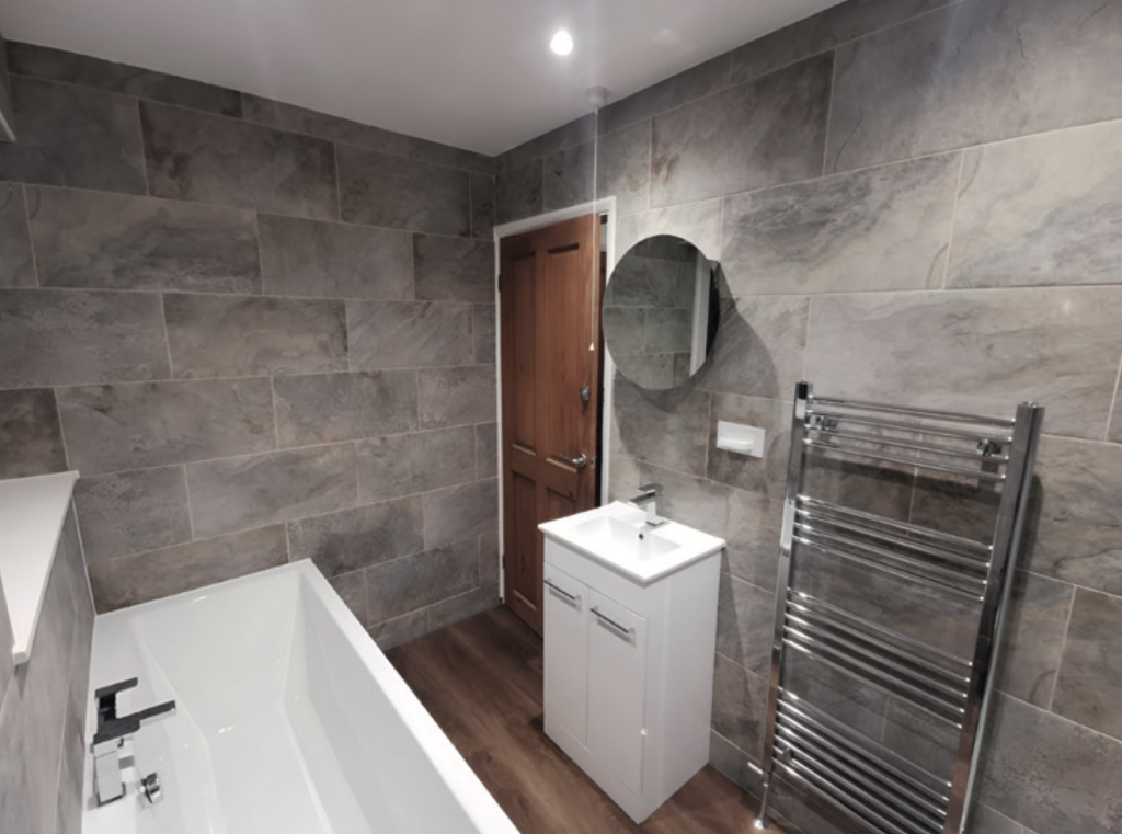 A modern bathroom with grey stone-effect tiled walls and a wood-look floor. On the left is a white bathtub with a black tap and fittings. In the centre, a small white vanity unit with a built-in sink sits beneath a round wall-mounted mirror. Behind it is a wooden door with a white frame. On the right, a chrome heated towel rail is fixed to the wall. The space is lit by a ceiling spotlight, giving the room a clean, contemporary appearance.