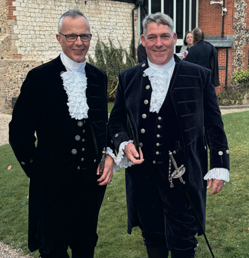 Jo Pearson stands alongside outgoing High Sheriff Patrick Lines, both wearing traditional ceremonial dress with lace jabots and dark coats, posing outdoors on a gravel path beside a lawn.