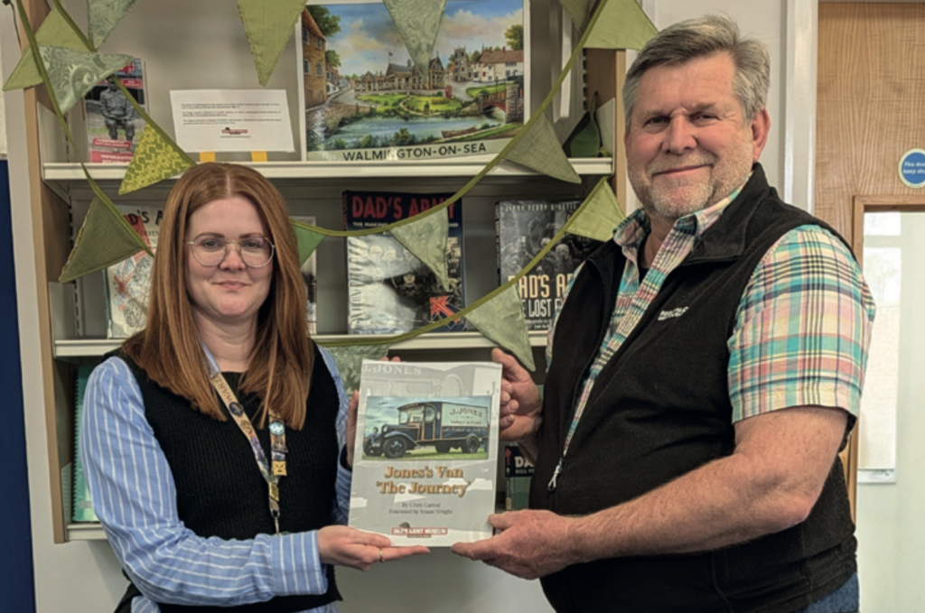 A man and woman stand indoors in front of a bookshelf display decorated with bunting, both holding a book titled Jones’s Van “The Journey”, which features an illustration of a vintage delivery van on the cover. The woman wears glasses and a lanyard, while the man is dressed in a checked shirt and dark fleece. Books and local history titles are visible behind them.