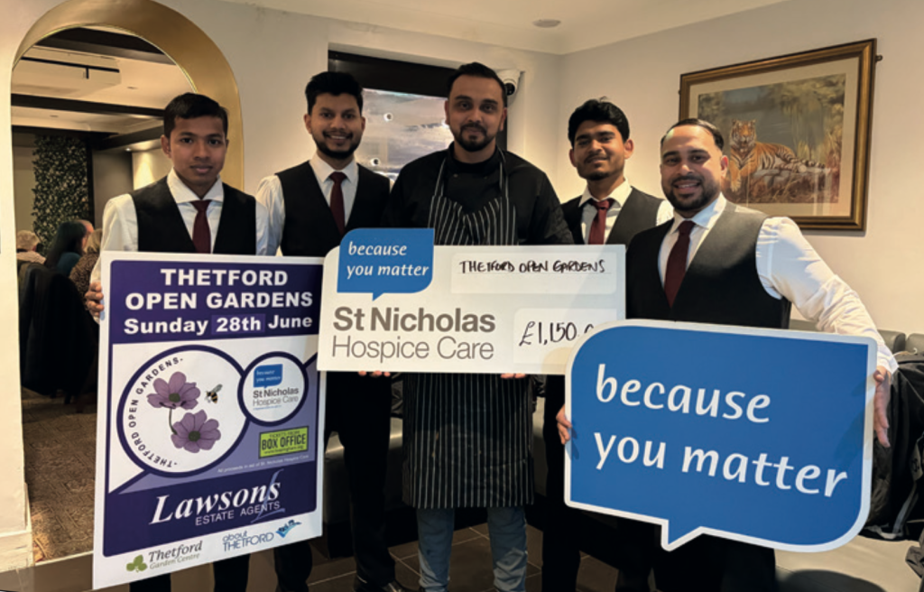 Group of New Saffron staff standing indoors holding a large cheque for £1,150 in support of St Nicholas Hospice Care, alongside promotional boards for Thetford Open Gardens.