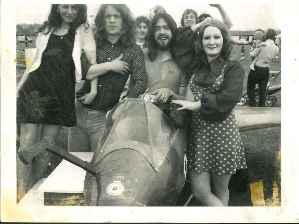 Vintage black-and-white photograph of a group of young adults gathered outdoors around a small aircraft cockpit or fuselage, smiling and posing for the camera. The group wear 1960s–1970s style clothing, including patterned dresses and long hair. In the background, other people, cars, and a pram are visible on a grassy field. The photo shows signs of age, with scratches, fading, and worn edges.