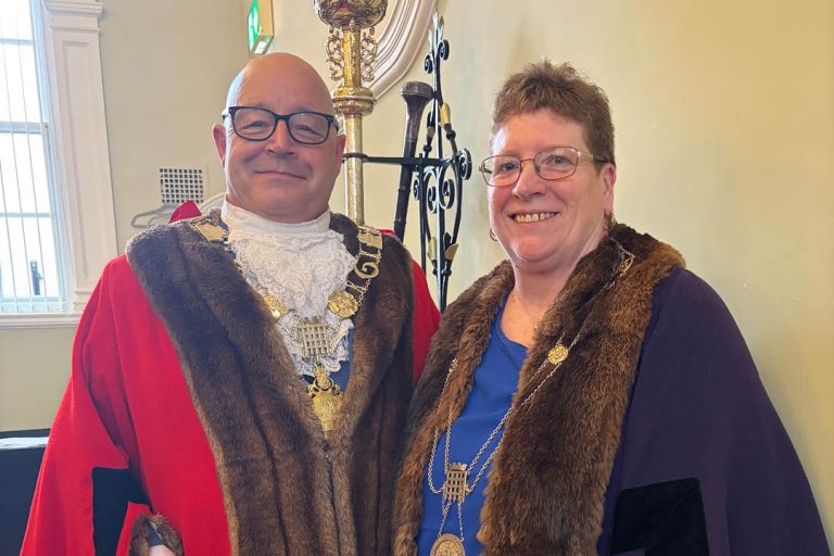 A man and woman wearing ceremonial mayoral robes and chains stand smiling together indoors. The man is dressed in a traditional red robe with fur trim and a large gold chain of office over a white lace cravat, while the woman wears a dark robe with fur trim and a gold chain over a blue top. Ornate interior details, including a decorative staff and staircase railing, can be seen in the background.