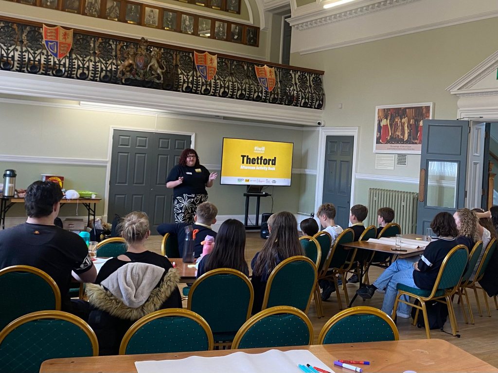 Young people sit at tables in a historic hall listening to a speaker giving a presentation beside a screen reading “Thetford Afternoon Activity Pack”. Pens and paper are laid out on the tables, while refreshments are visible at the back of the room.