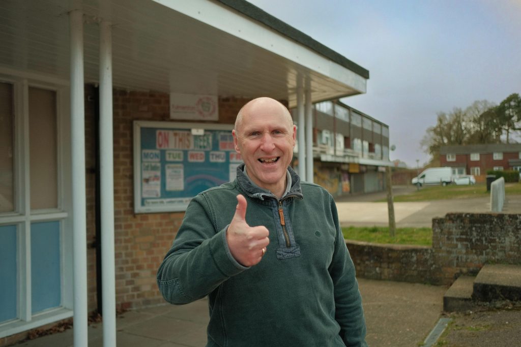 A man gives a thumbs up while standing outside a building and parade of shops on an overcast day.
