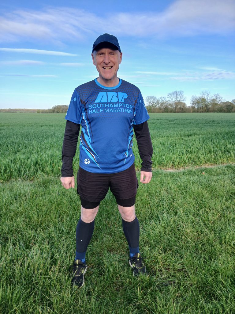 A man wearing running gear and a blue Southampton Half Marathon shirt stands smiling in a grassy field under a blue sky.