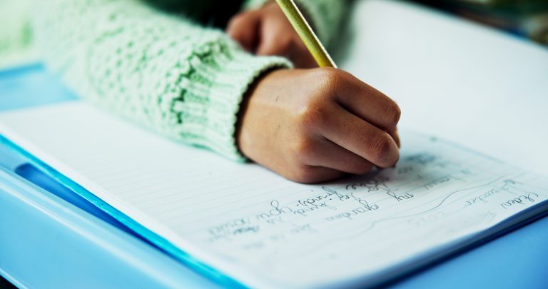 Close-up of a child’s hand writing in an exercise book with a pencil. The child is wearing a pale green knitted jumper, while handwritten notes and a blue folder are visible on the desk.