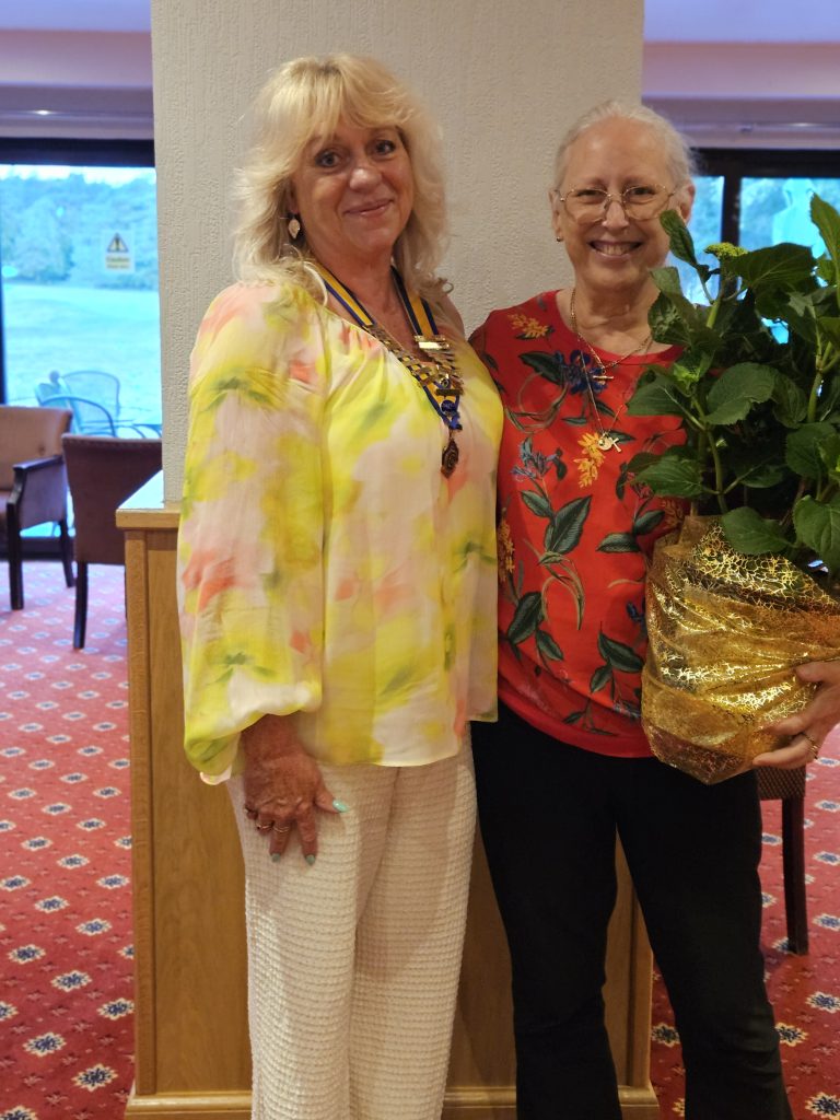 Two women stand smiling together indoors during an Inner Wheel handover. One wears the ceremonial chain of office, while the other holds a wrapped hydrangea plant presented in recognition of her service. The image marks the transition to a new club president and celebrates the outgoing president's contribution.