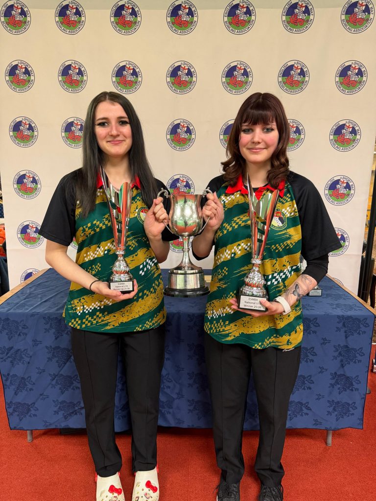 Two young bowlers pose with trophies and a silver cup in front of an English Short Mat Bowling Association backdrop after a competition.