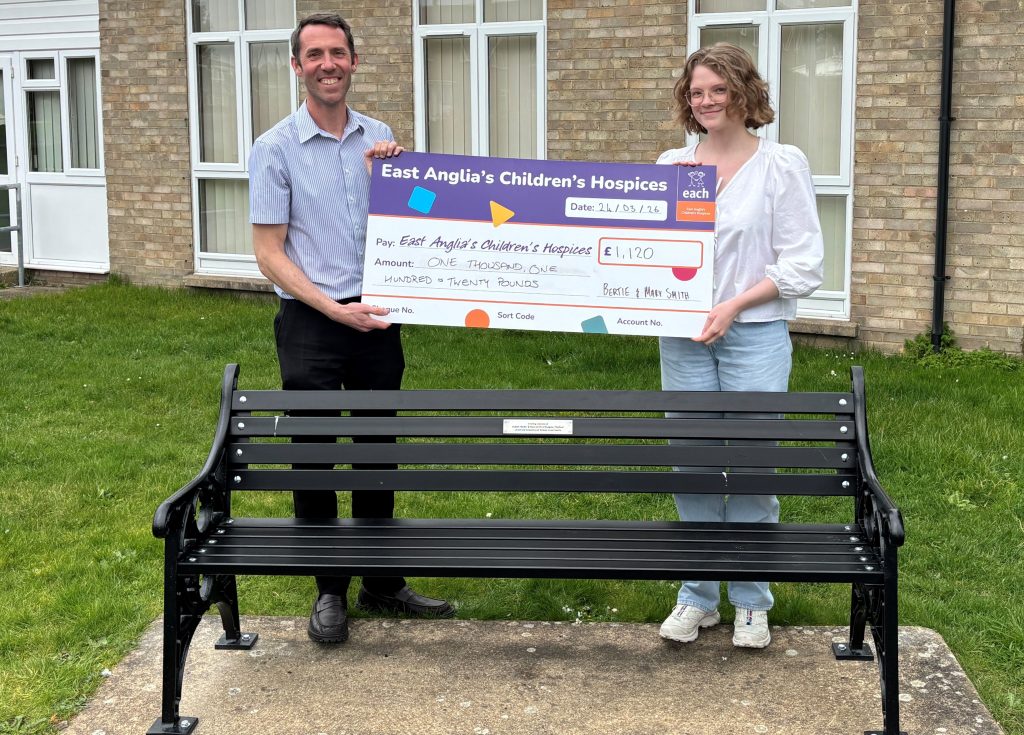 EACH Media and PR Manager Matt Plummer and Senior Digital Communications Coordinator Grace McGill stand outside holding a large presentation cheque for £1,120 made payable to East Anglia’s Children’s Hospices in memory of Bertie and Mary Smith. A black memorial bench is positioned in the foreground.