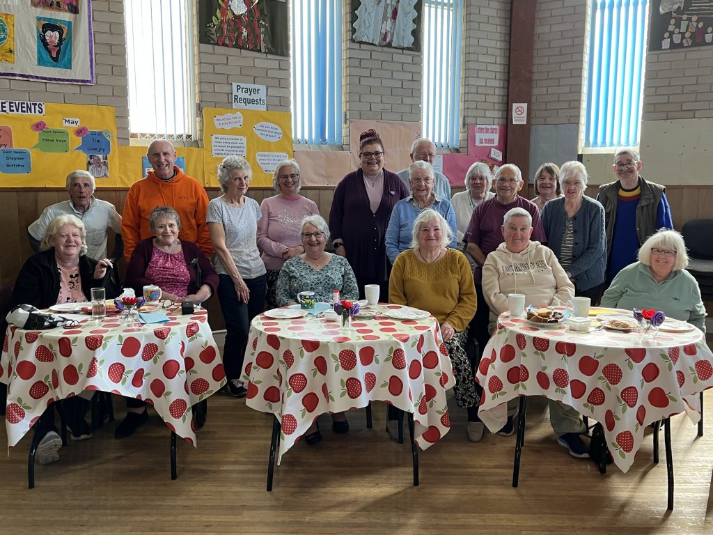 A group of older residents and organisers pose together inside a community hall during a social gathering. Several people are seated at tables covered with apple-patterned tablecloths, with cups, plates and refreshments in front of them. Colourful noticeboards and signs including “Prayer Requests” and upcoming events are displayed on the walls behind the group.