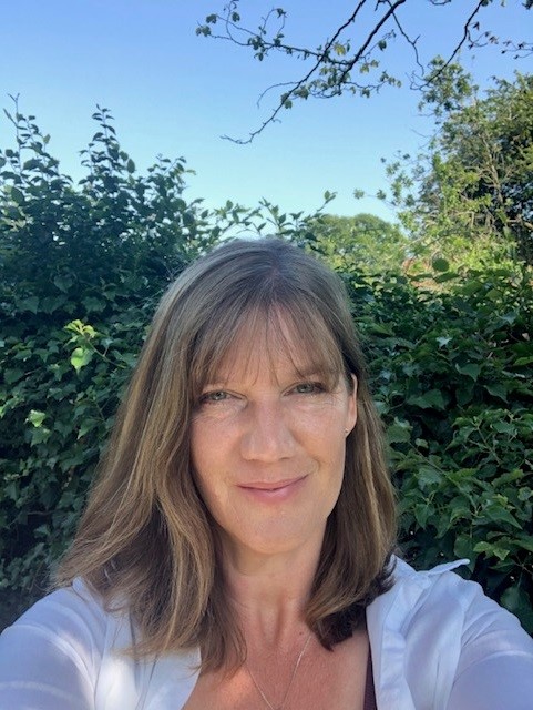 Portrait photograph of a woman outdoors standing in front of green hedges and trees under a bright blue sky. She has shoulder-length brown hair and is wearing a white top and necklace, smiling gently towards the camera.