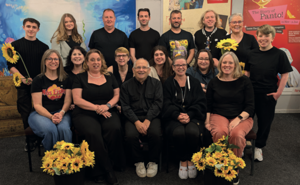 Group cast photo of Thetford Players members posing indoors with sunflower decorations for Calendar Girls The Musical.