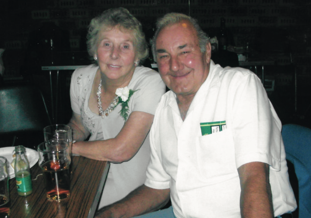 An older couple, Bertie and Mary Smith, sit together smiling at a social event. Mary wears a light-coloured dress with a corsage and necklace, while Bertie wears a white short-sleeved shirt. Drinks and glasses sit on the table in front of them.