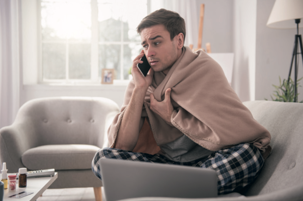 A man wrapped in a beige blanket sits on a sofa at home while talking on a mobile phone, looking unwell and concerned. He is wearing checked pyjama trousers and has a laptop open in front of him, with medicine bottles and tissues on a nearby table. Bright daylight streams through the windows behind him, and the living room includes pale furniture and a floor lamp.