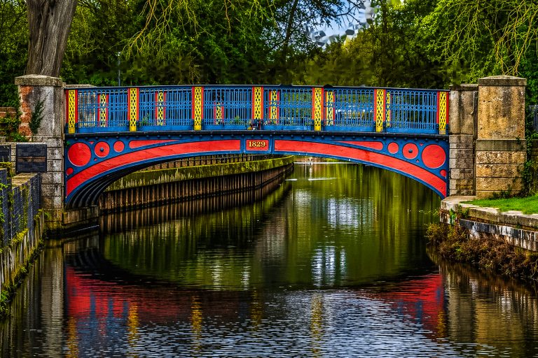 Photo of the brightly coloured iron bridge in Thetford, painted blue, red and yellow, spanning a calm river or canal. The bridge features the year “1829” at its centre, with its vivid colours reflected in the water below. Trees and greenery surround the scene, while stone pillars stand at either end of the bridge.