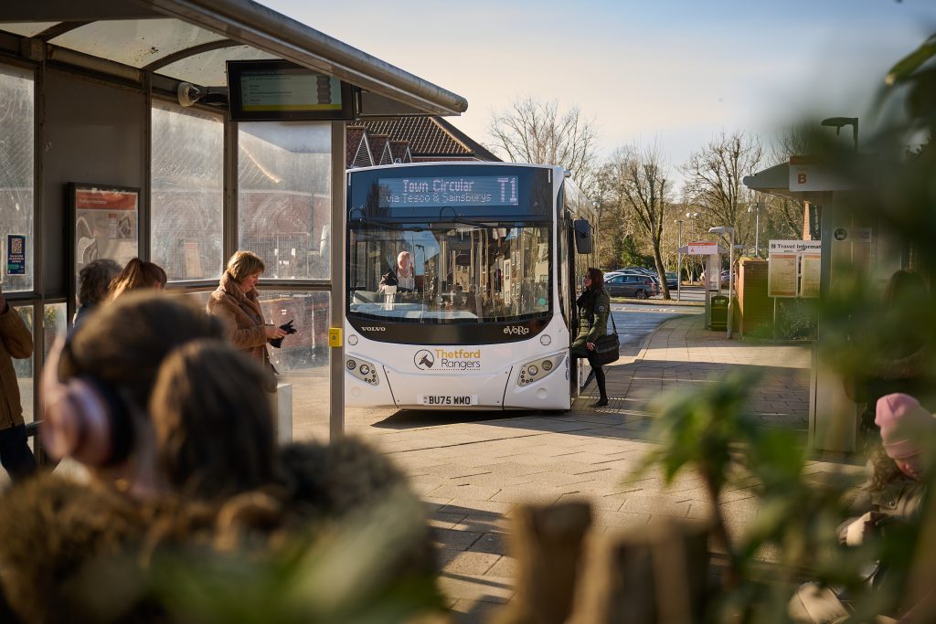Photo of a Thetford Rangers T1 bus waiting at a bus stop in Thetford town centre on a sunny day. The digital destination display reads “Town Circular T1 via Tesco & Sainsburys”. Several people are waiting at the bus stop or boarding the bus, while travel information boards and shelters are visible around the interchange. Trees and nearby buildings can be seen in the background, with blurred plants and passengers framing the foreground of the image.
