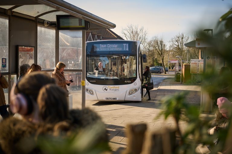 Photo of a Thetford Rangers T1 bus waiting at a bus stop in Thetford town centre on a sunny day. The digital destination display reads “Town Circular T1 via Tesco & Sainsburys”. Several people are waiting at the bus stop or boarding the bus, while travel information boards and shelters are visible around the interchange. Trees and nearby buildings can be seen in the background, with blurred plants and passengers framing the foreground of the image.