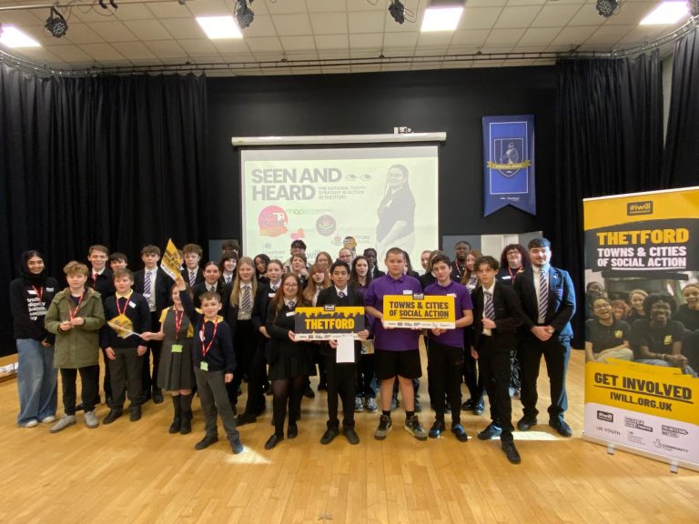 A large group of young people and organisers pose on a stage during a Thetford social action event. Several hold yellow signs reading “Thetford” and “Towns & Cities of Social Action”, while banners for the iWill campaign stand at the side of the stage. A screen behind them reads “Seen and Heard - The National Youth Strategy in Action in Thetford”.
