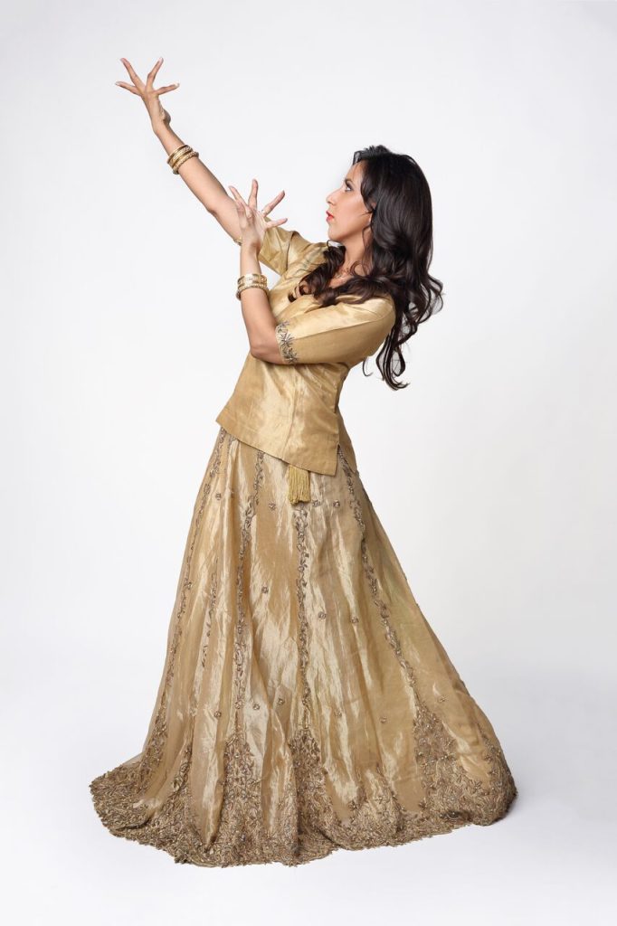 A studio portrait of a dancer wearing a gold embroidered traditional outfit with matching bangles. She strikes an expressive dance pose with both arms raised and fingers elegantly positioned against a plain white background.