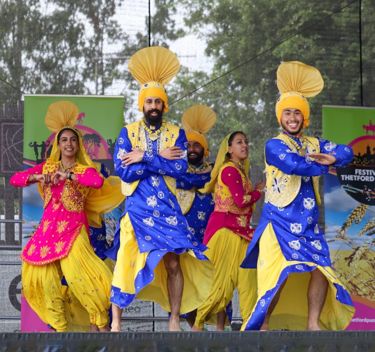 A group of dancers in bright traditional Punjabi outfits perform on an outdoor stage during a festival event. The performers wear vivid blue and yellow costumes with matching turbans and pink accents, posing mid-routine with crossed arms and raised knees. Festival banners stand behind them, with trees visible in the background.