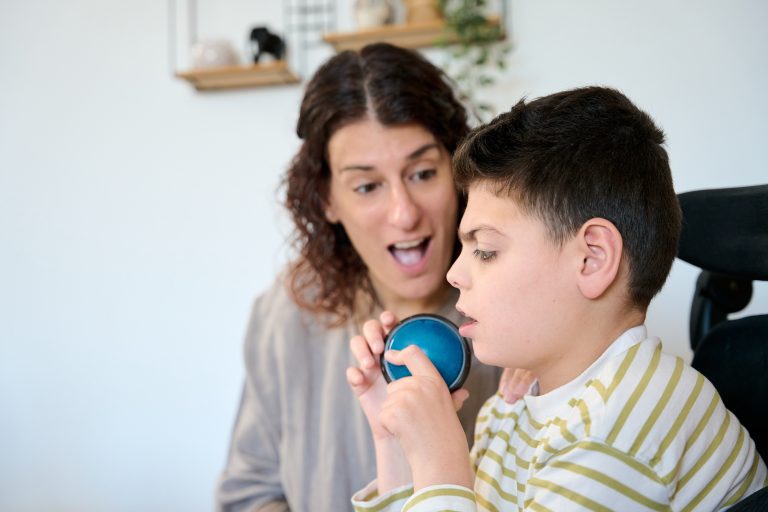 A young person uses a communication aid while seated indoors, with a supportive adult looking on attentively.