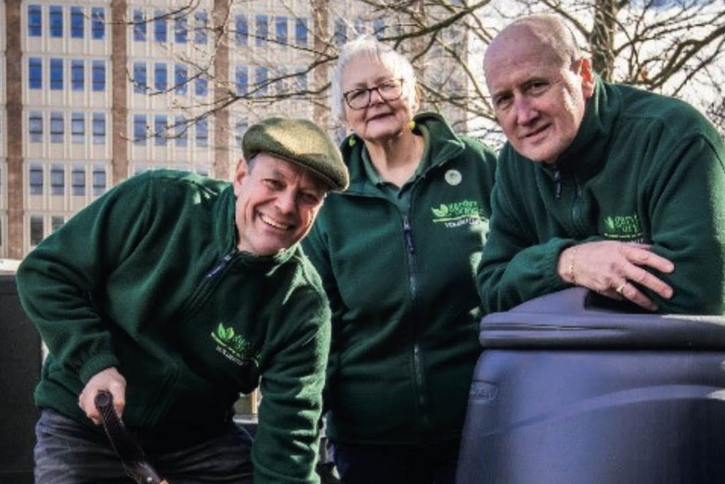 Three Master Composter volunteers stand beside a compost bin, with one holding a garden fork, promoting home composting and waste reduction.