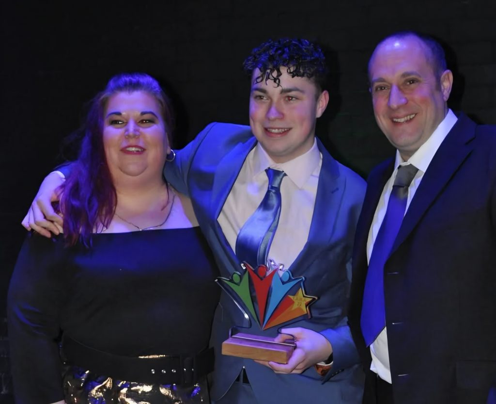 Three people pose together at an awards ceremony, smiling towards the camera against a dark stage backdrop lit with blue and purple lighting. The person in the centre wears a light blue suit and tie and holds a colourful trophy featuring stylised figures and a star. The two people beside them are dressed in formal evening attire, with one person’s arm around the award winner. The image captures a celebratory moment following a presentation or recognition event.